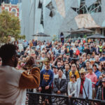 a man plays trumpet on stage at fed square in front of a huge crowd of people
