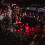 a room filled with red light, plants and people playing an electric bass, various keyboards and drum pad. there's a large audience looking on.