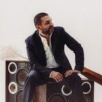a man in a suit sits on retro speakers, he glances to the side with hand on knee