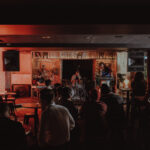 a darkened room with a warm red glow, there's an artist performing on stage and a dimly lit crowd
