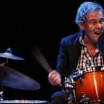 a smiling man in a check shirt plays the drums