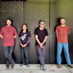 four people in casual cloths stand smiling in front of a metal walll