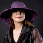 a red lipsticked woman in a black silk shirt and purple hat gently smiles at the camera