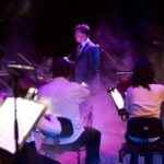 On a stage bathed in haze and pink light, a man in a suit stands centred, surrounded by seated musicians in white shirts.