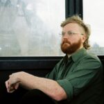 Man with glasses and a crisp green shift sits at a window (maybe on a train) looking ponderous