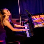Liv sits at a black piano on stage, head back in song